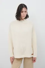 Polera de tejido de punto en color crema, con cuello alto, diseño holgado y costura central vertical visible. Presenta mangas largas con puños acanalados y una pequeña etiqueta decorativa en el ruedo.