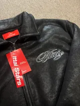 Campera de cuero sintético negro con diseño tipo bomber, cuello camisero y puños y ruedo elásticos. Presenta un bordado grande en la espalda con tipografía cursiva en color blanco.