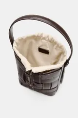 Bolso tipo bucket de diseño trenzado en color marrón oscuro con costuras a la vista en contraste. Cuenta con asa de mano fija, correa bandolera extraíble y compartimento interior de tela en color crudo con cierre mediante fruncido.
