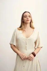 Vestido largo color beige claro, con escote en V, manga corta acampanada y cierre frontal de botones. Presenta un lazo ajustable en la cintura y un tejido con textura sutil.