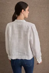 Blusa de lino color blanco con cuello mao, cierre frontal con botones, mangas largas abullonadas con puños ajustados y detalle de volado en el ruedo.