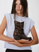 Botas estilo cowboy de cuero en tono marrón con efecto desgastado. Presentan un diseño de caña media con detalles de correas, argollas metálicas y tachas decorativas. Cuentan con un taco cuadrado de altura media y punta ligeramente cuadrada.
