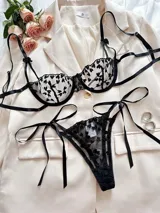 Conjunto de lencería negro de dos piezas con bordado de corazones y detalles de tiras con lazos. Incluye un sostén con aros y tirantes regulables, y una bombacha colaless ajustable con lazos en los laterales.