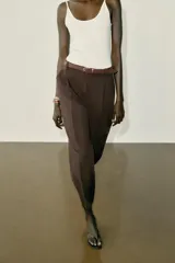 Pantalón tobillero de tiro medio, color beige, con pinzas delanteras y bolsillos frontales. Incluye un cinturón fino de cuero marrón con hebilla metálica dorada y cierre frontal con cremallera, botón interior y gancho.