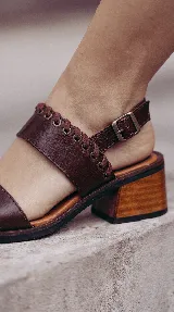 Sandalia de cuero color marrón, con taco bajo de madera, tiras anchas en el empeine y detalle de cordones entrelazados en el lateral. Cuenta con tira ajustable con hebilla en el talón.