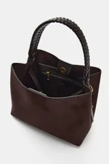 Bolso de mano de formato saca, confeccionado en material tipo gamuza color marrón oscuro, con asa superior trenzada en color negro.