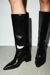Bota alta de caña media con acabado brillante, tacón cuadrado y punta fina. Diseño minimalista con costura vertical lateral y estilo pull-on.