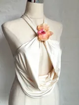 Top color crema o amarillo pálido, estilo halter con cuello atado y escote pronunciado en V con drapeado frontal. El cuello está adornado con una flor de orquídea en tonos amarillo, naranja y rosa.