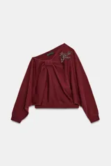 Jersey de punto color bordó con cuello asimétrico, detalle de nudo en el escote y pedrería en forma de hoja en el hombro. Manga larga.