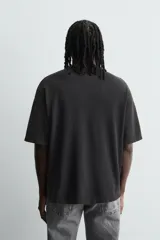 Camiseta de punto negro con efecto lavado, corte relaxed fit y cuello redondo. Presenta un estampado frontal con tipografía gótica en azul y una imagen circular que simula un iris en tonos rojizos y amarillos.