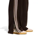 Pantalón Adidas Firebird Loose de corte holgado y diseño deportivo, color marrón con las tres tiras características de la marca en los laterales y el logo del trifolio bordado en color rosado claro. Cuenta con cintura elástica y bolsillos laterales con cierre.