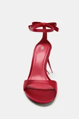 Sandalia de tacón alto fino, color rojo satinado, con tira delantera y cierre mediante tira con hebilla a la pulsera del tobillo con detalle de lazo trasero. Acabado en punta.
