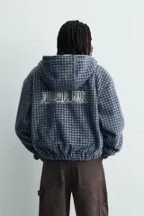 Cazadora cropped de corte holgado confeccionada en tejido texturizado con estampado de cuadros. Presenta cierre frontal con cremallera, capucha, mangas largas con puños elásticos y bolsillos frontales tipo canguro.