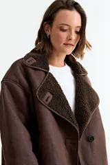 Chaqueta de estilo aviador en color marrón, confeccionada en material simil gamuza con interior y solapas de corderito sintético. Presenta cierre frontal con botones, corte holgado y mangas largas.