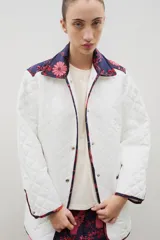 Campera acolchada de corte holgado, color blanco con diseño capitoneado en forma de rombos. Presenta cuello camisero y bordes internos con estampado floral en tonos azul y rojo, cierre frontal con botones a presión y bolsillos laterales.