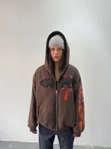 Campera reversible con cierre frontal. Un lado confeccionado en algodón color marrón con estampa plastisol en color naranja, bordados con apliques y capucha. El otro lado confeccionado en tela polar de pelo largo color negro con capucha.