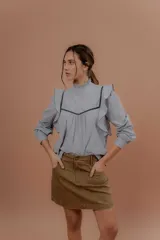 Blusa de jeans fino color celeste con detalles de terciopelo negro en contraste. Presenta cuello alto con volados, mangas largas abullonadas con puños ajustados y volados decorativos en los hombros y el frente.