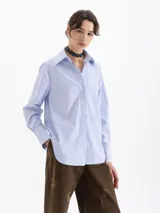 Camisa de poplin a rayas verticales celestes y blancas, con cuello clásico, cierre frontal de botones y mangas largas con puños abotonados. Presenta un sutil bordado de llamas distribuido por toda la prenda.