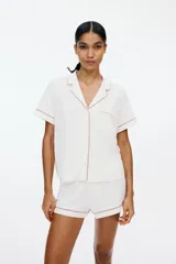 Pijama de dos piezas confeccionado en mezcla de viscosa, compuesto por una camisa de manga corta con cuello abierto, botones frontales y bolsillo superior, y un short con elástico en la cintura. Ambos presentan ribetes decorativos en contraste.