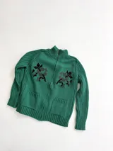 Cardigan de punto color marrón con cierre frontal, cuello alto y bolsillos aplicados. Presenta diseño tejido localizado con detalles en violeta y negro en el pecho.