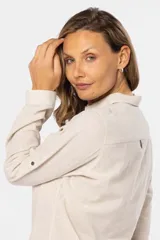 Camisa blanca de manga larga con cuello y tapeta de medio botón. Presenta dos bolsillos frontales con tapa y botones metálicos. Las mangas tienen presillas para sujetar el puño enrollado.