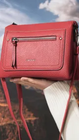 Bandolera de tamaño mediano en color gris, confeccionada en símil cuero con textura granulada. Presenta un bolsillo frontal con cierre, herrajes metálicos oscuros y correa ajustable y desmontable.