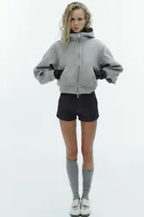 Cazadora de estilo deportivo con capucha ajustable, cierre frontal de doble cremallera y mangas abullonadas con puños elásticos. Presenta bolsillos delanteros y bajo elástico.