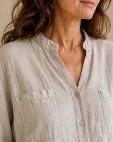 Camisa de lino color beige, de corte holgado, con cuello mao, bolsillos frontales en el pecho, mangas largas remangables y cierre frontal con botones.