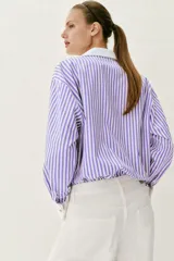 Camisa de popelín con diseño de rayas verticales en tonos blanco y lila, cuello solapa con escote en pico, manga larga y puños anchos contrastantes en color blanco. Cierre frontal con botones metálicos dorados.