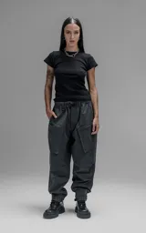 Pantalón de estilo utilitario confeccionado en textil técnico ripstop color gris oscuro. Presenta un corte oversize con cintura elástica, cierre frontal con broche técnico, bolsillos delanteros de gran tamaño con cierre y sistema de ajuste en los tobillos mediante broches snap.