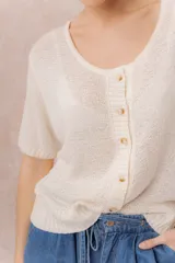 Cardigan tejido de manga corta color beige, con escote redondo amplio y abotonadura frontal con botones al tono. Presenta un calce relajado y terminaciones en rib en la cartera y el ruedo.