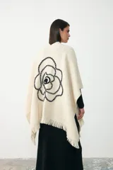 Ruana de tejido artesanal en color crudo, con diseño de rosa bordada en relieve en color negro. Presenta terminaciones con flecos en los bordes.