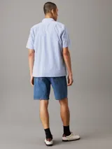 Camisa de manga corta Calvin Klein confeccionada en popelín de algodón elástico con diseño de rayas verticales en blanco y celeste. Presenta cuello en punta, cierre frontal de botones, bolsillo de parche en el pecho y bajo recto.