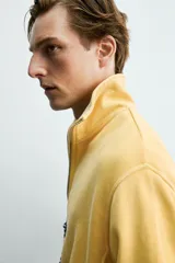 Sudadera de corte relajado confeccionada en algodón con interior de rizo, color amarillo mostaza con efecto lavado. Presenta cuello subido con cierre frontal de cremallera y detalle de cinta trenzada en el tirador, manga larga y acabados en rib.