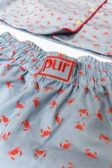 Pijama de dos piezas, compuesto por una camisa de manga corta con cuello solapa y botones frontales, y un short a juego. El conjunto es de color azul claro con un estampado repetitivo de pequeños motivos florales en color rosado/rojo, y ribetes de color rojo en los bordes de la camisa y el short.