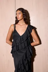 Vestido largo negro con escote en V y tirantes finos. Presenta volados verticales en cascada a lo largo del cuerpo y un tajo pronunciado en la parte delantera de la falda. La espalda es baja con tiras ajustables.