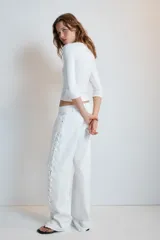 Jeans rectos de tiro medio en color blanco, con cinco bolsillos, trabillas y cierre frontal con cremallera. Presenta detalle de cordones cruzados con ojalillos metálicos a lo largo de los laterales de ambas piernas.
