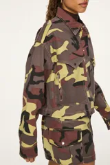 Chaqueta de denim con estampado camuflado en tonos marrón, verde y beige, de corte recto y largo recortado. Cuenta con cuello camisero, cierre frontal con cremallera, bolsillos con solapa en el pecho y solapas decorativas en los hombros.