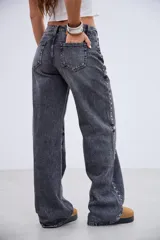 Jean de tiro medio en color negro lavado, con corte recto y amplio. Presenta detalles de tachas metálicas a lo largo de las costuras laterales y en la zona de los bolsillos delanteros.