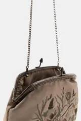 Bolso pequeño tipo cartera con cierre de clip metálico, confeccionado en tela color beige con bordados florales en tonos tierra y verdes. Cuenta con una fina cadena metálica para llevar al hombro y forro interior.
