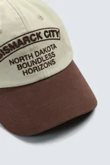 Gorra de seis paneles de algodón en color beige con visera marrón contrastante. Cuenta con un diseño bordado en marrón en el frente con texto alusivo.