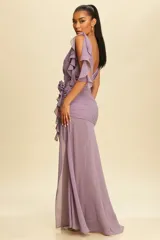 Vestido largo de fiesta color lila, con escote en V, mangas cortas con volados, detalle de frunces en la cintura y una flor decorativa lateral. Presenta una falda con tajo frontal y caída fluida.