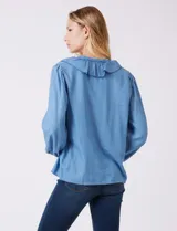 Blusa de lyocell color azul claro, con escote en V y volados en el cuello y la parte frontal. Tiene mangas largas con puños elásticos y cierre de botones en la parte delantera.