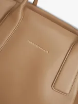 Cartera tote Tommy Hilfiger de color tostado, con acabado liso, dos asas superiores y cierre de cremallera. Cuenta con un compartimento principal, un bolsillo interior con cremallera y el logo metálico de la marca en relieve en la parte delantera.