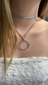 Gargantilla tipo choker de gamuza gris, compuesta por una tira doble que rodea el cuello y un dije circular metálico plateado suspendido de una tira central.