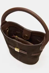 Bolso tipo saca de tamaño mini, confeccionado en gamuza color marrón oscuro con detalles en cuero marrón. Presenta un asa de mano fija, una tira decorativa horizontal con hebilla metálica dorada en el frente y costuras verticales visibles.