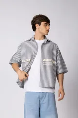 Camisa de manga corta de corte boxy, confeccionada en popelina a rayas celestes y blancas. Presenta cuello camisero clásico, cierre frontal con botones y estampado tipográfico en el pecho.