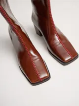 Botas de caña media color marrón, confeccionadas en cuero con acabado brillante. Presentan punta cuadrada, taco de madera y un distintivo bordado decorativo en color rojo que recorre el empeine hasta la punta.