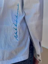 Camisa de algodón a rayas blancas y azules, con cuello sastrero, solapa, cartera frontal con botones de nácar y bolsillo aplicado en el delantero con bordado. En la espalda incorpora bordado distintivo de la marca.