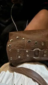 Cartera de hombro color marrón con acabado texturizado, solapa frontal decorada con tachas metálicas plateadas y una argolla central. Cuenta con correa ajustable y cierre principal.
